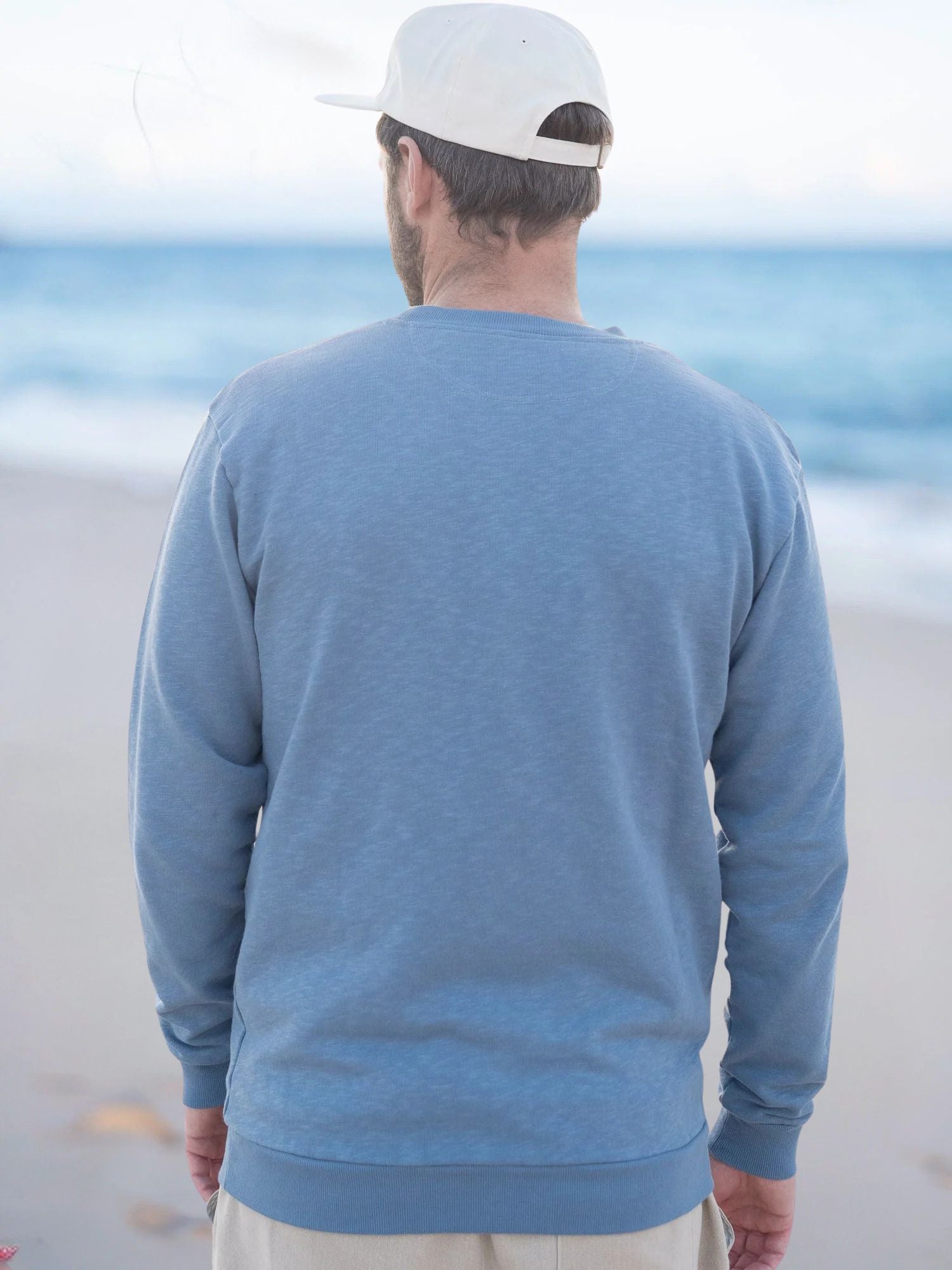 Ein junger Männer trägt einen blau melierten Fair Fashion Pullover am Strand in Portugal aus 100% GOTS zertifizierter Biobaumwolle. Ansichten von hinten.