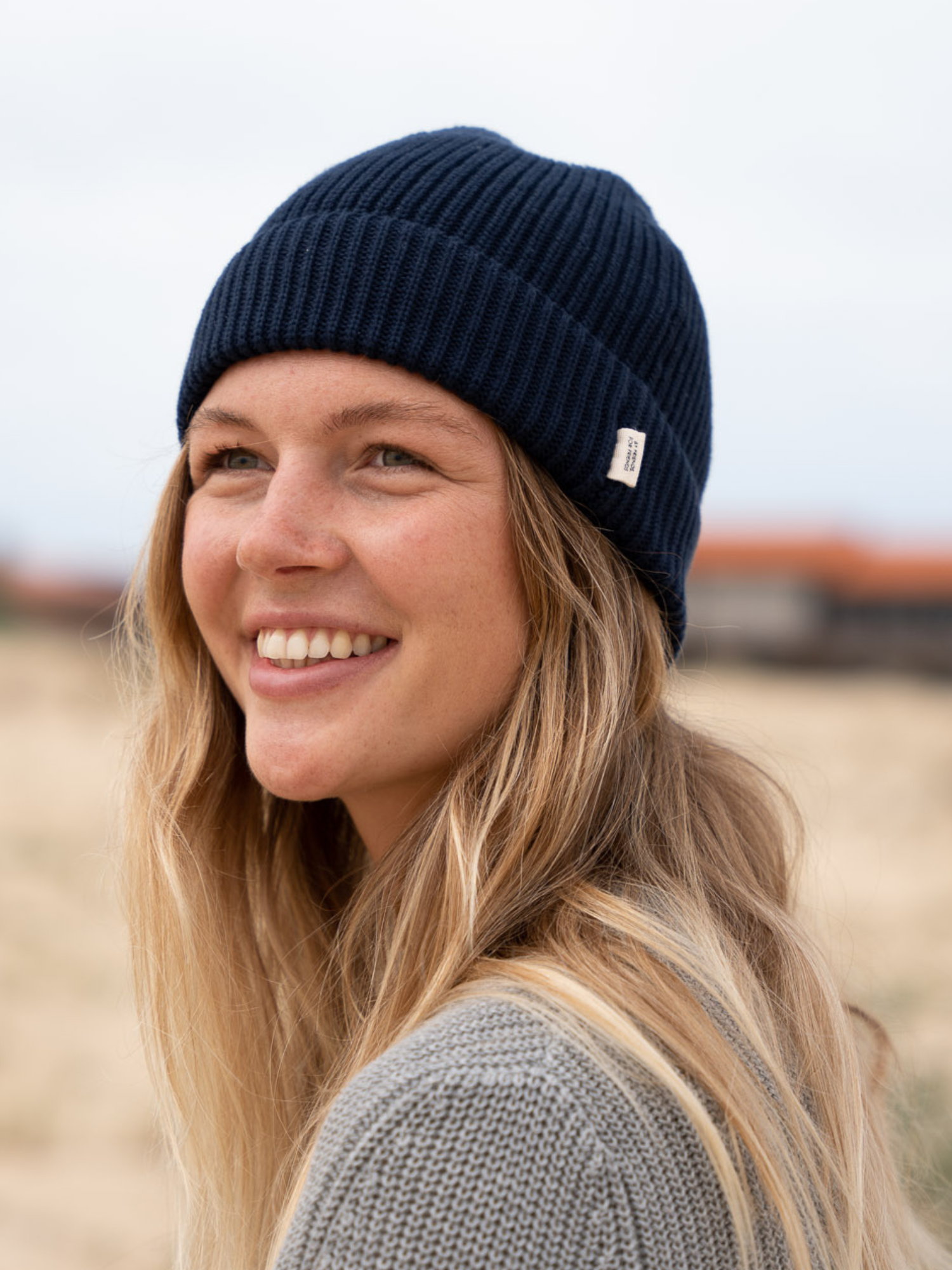 Eine junge Frau steht am Strand und trägt eine vegane Strickmütze in Navy. Sie lächelt an der Kamera vorbei und schaut in den Himmel.