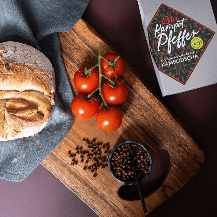 Conflictfood Kampot Pfeffer Rot Verpackung, daneben ein Brett mit Brot, Tomaten und Pfefferkörnern in einer Schale