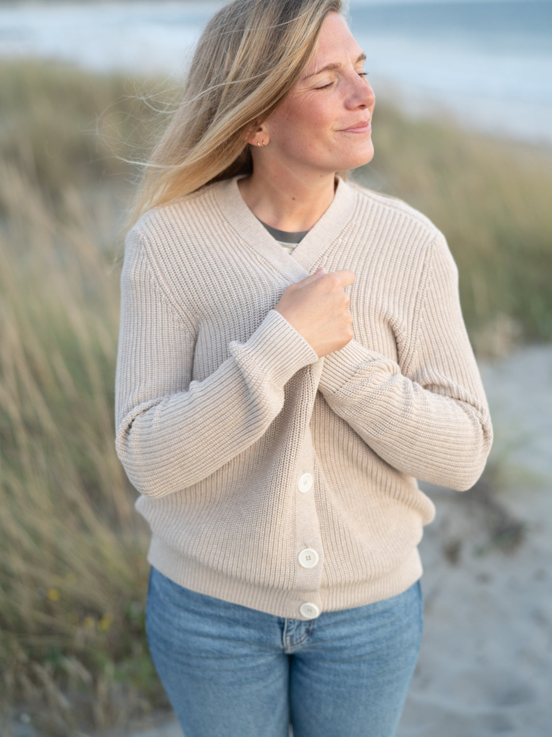 FUXBAU 4Seasons Frauen Strickjacke in beigemeliert aus 100% GOTS zertifizierter Biobaumwolle. Gefertigt in Italien und gertragen von einer jungen Frau am Strand in Portugal.