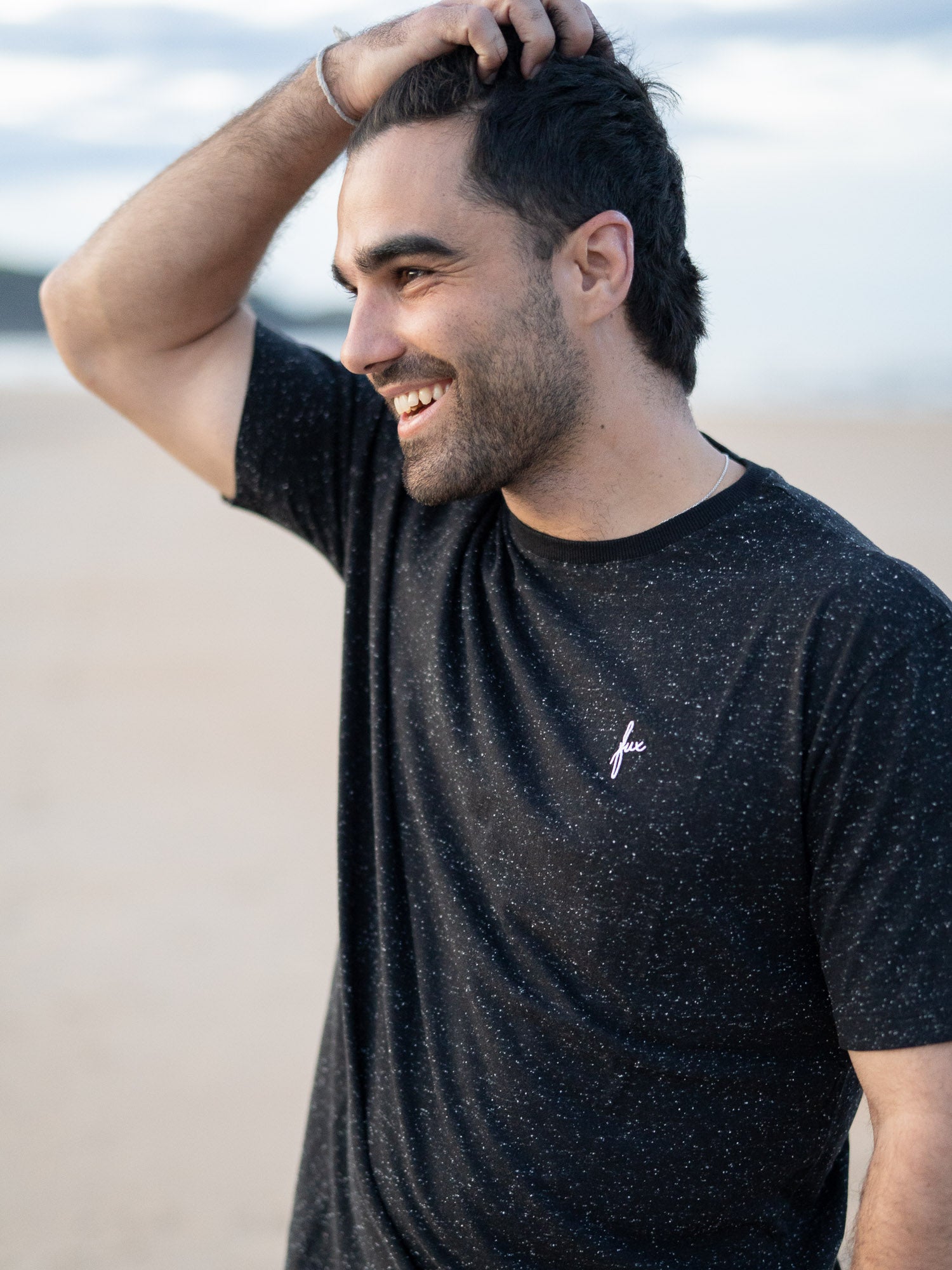 Ein junger Mann trägt ein schwarzes T-Shirt mit kleinen, weißen Punkten von FUXBAU aus Biobaumwolle am Strand in Spanien.