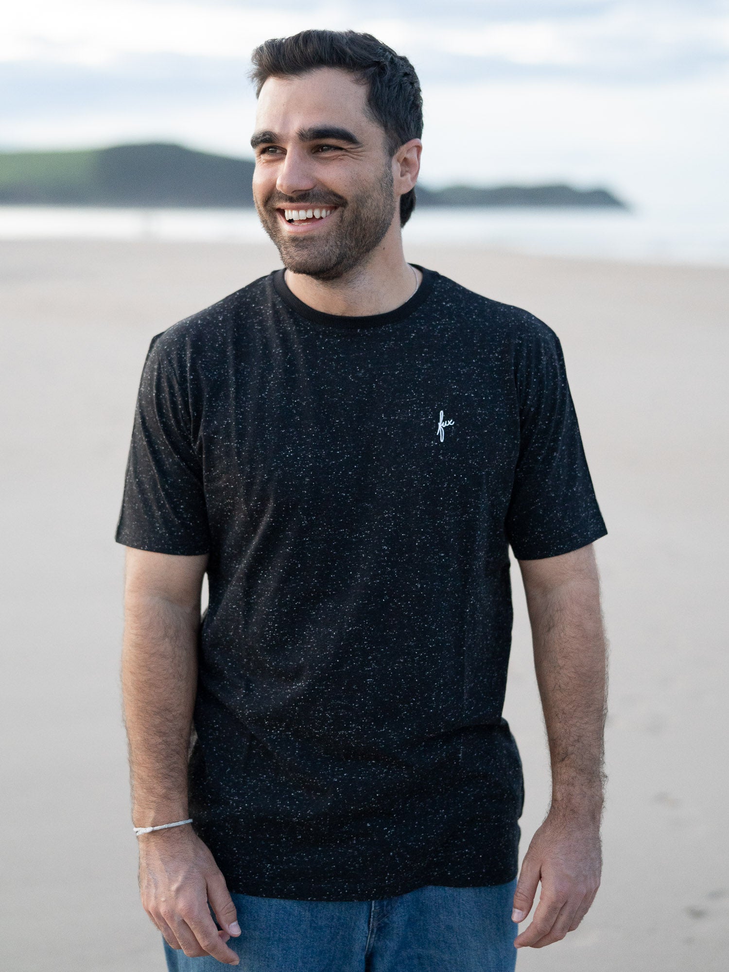 Ein junger Mann trägt ein schwarzes T-Shirt mit kleinen, weißen Punkten von FUXBAU aus Biobaumwolle am Strand in Spanien.