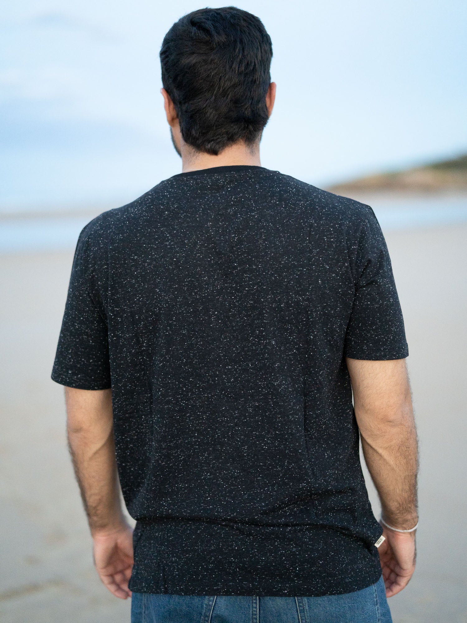 Ein junger Mann trägt ein schwarzes T-Shirt mit kleinen, weißen Punkten von FUXBAU aus Biobaumwolle am Strand in Spanien.